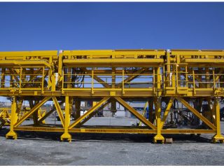 Large, bright yellow industrial steel structure with railings, platforms, and support beams, standing on gravel outdoors under a clear blue sky.