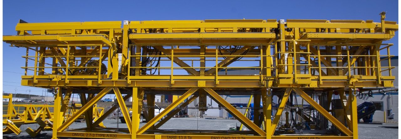 Large yellow industrial steel structure with platforms, railings, and ladders, standing on a gravel surface under a clear blue sky. The structure appears to be used for construction or heavy lifting.