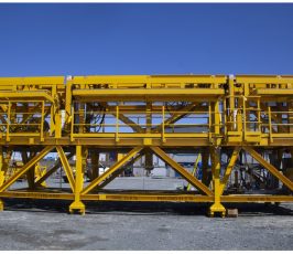 Large yellow industrial steel structure with platforms, railings, and ladders, standing on a gravel surface under a clear blue sky. The structure appears to be used for construction or heavy lifting.