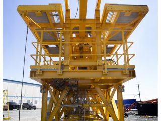 A large, bright yellow industrial structure with platforms, railings, and cables stands outdoors on gravel near a warehouse under a clear sky.