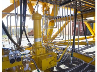 Close-up view of intricate metal pipes and hydraulic components on a large yellow industrial machine, with sunlight casting shadows and a fence and container visible in the background.
