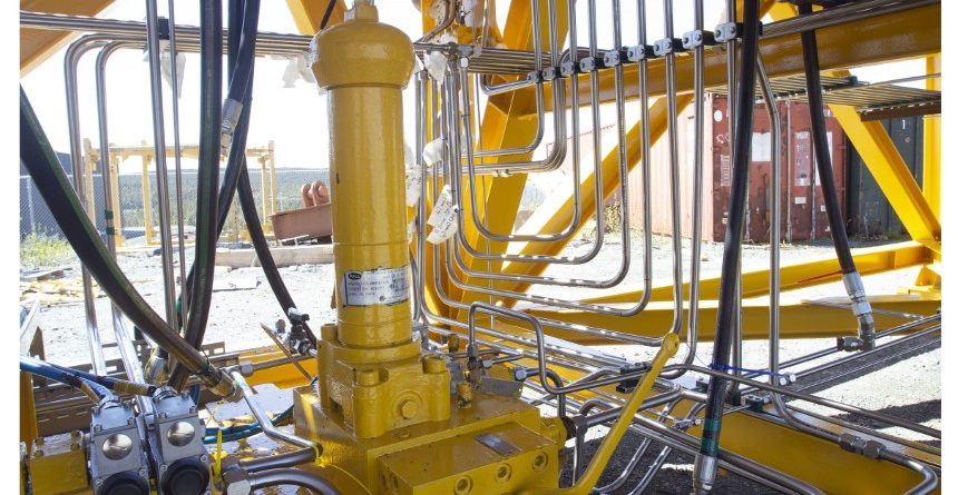 Close-up view of intricate metal pipes and hydraulic components on a large yellow industrial machine, with sunlight casting shadows and a fence and container visible in the background.