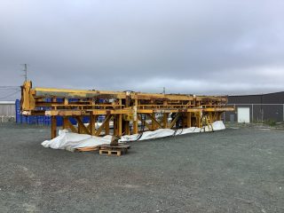 A large yellow industrial structure is positioned on a gravel lot, with some white tarp and wooden pallets nearby. The sky is overcast, and a warehouse building is visible in the background.