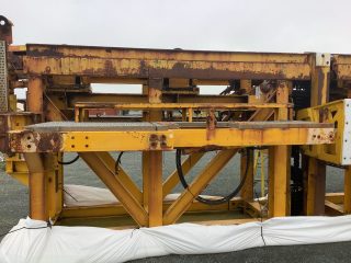 A large, yellow, metal industrial structure with significant rust and corrosion stands on a gravel surface. White plastic sheeting is wrapped around part of the base. The sky is overcast.