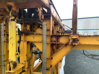 A close-up of a large, rusted yellow metal structure outdoors on gravel, with parts labeled in black marker. An industrial building is visible in the background.
