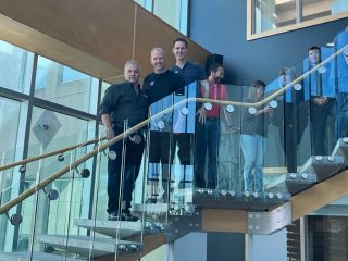 A group of people stand and pose for a photo on a glass and wood staircase inside a modern building with large windows and natural light. Three people are at the center, with others partially visible on the stairs.