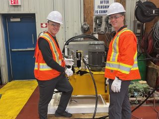 Two people wearing white hard hats, safety glasses, and orange reflective vests stand in an industrial setting beside machinery. They are smiling at the camera. The area has safety signs and industrial equipment in the background.