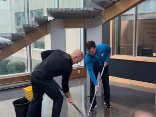 Two people with mops clean a shiny floor together in a modern building, near a staircase with glass railings and a yellow mop bucket. Large windows let in natural light.
