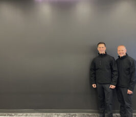 Two men wearing black jackets and pants stand side by side, smiling, against a large plain gray wall in a room with a patterned floor.