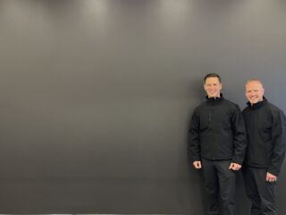Two men wearing black jackets and pants stand side by side, smiling, against a large plain gray wall in a room with a patterned floor.