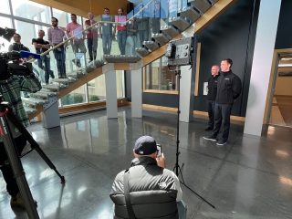 Two people stand in front of a camera for an interview in a modern building lobby, with a group of people watching from a glass staircase above. A photographer kneels in the foreground, and studio lights are set up.