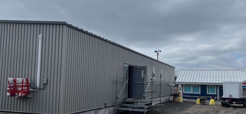 A gray metal building with a staircase and scaffolding at the entrance stands on a dirt lot under a cloudy sky. Yellow traffic cones and another building are visible in the background.