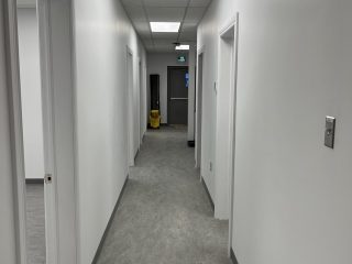A clean, empty hallway with white walls and closed doors on either side. The floor is gray, and fluorescent lights line the ceiling. At the end of the hall, a yellow cleaning cart is visible near a closed gray door.