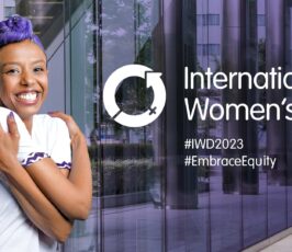 A smiling woman with purple hair wraps her arms around herself in a self-hug. Text reads “International Women’s Day,” with hashtags #IWD2023 and #EmbraceEquity on a purple-toned city background.