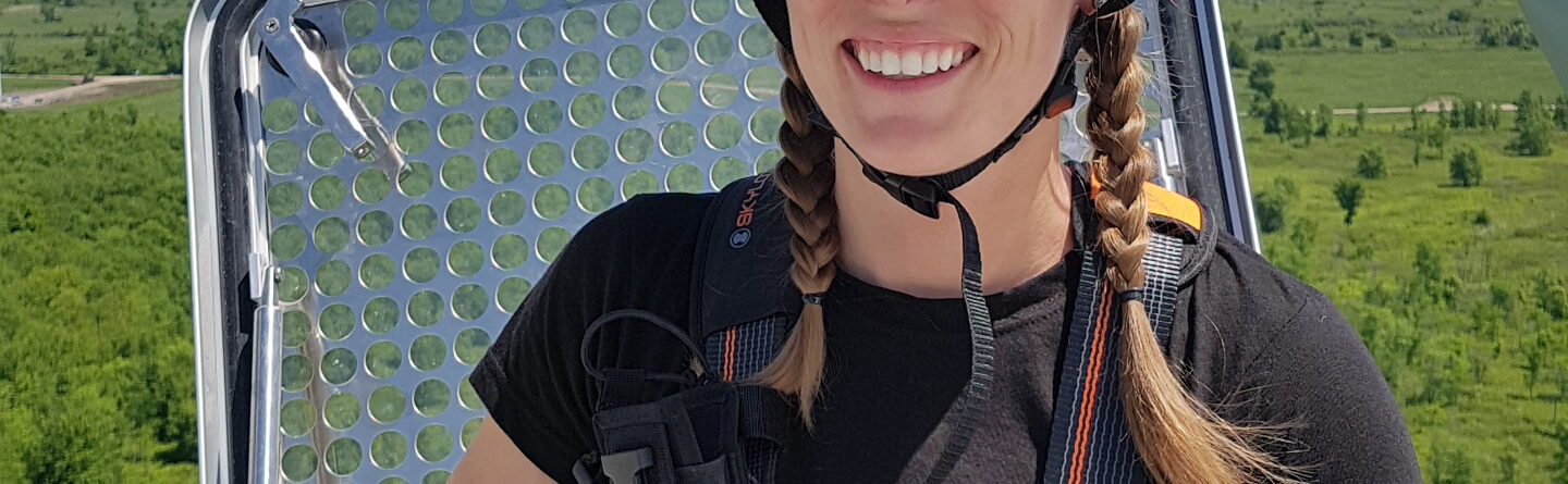 A smiling woman in safety gear, including a helmet and harness, stands at the top of a wind turbine with a scenic green landscape and blue sky in the background.