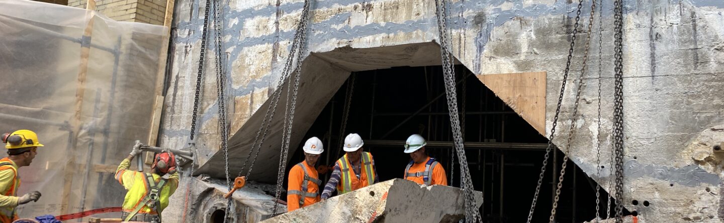 Construction workers in safety gear stand in front of a large opening in a concrete wall, surrounded by heavy machinery, chains, and cut concrete sections at a construction site.