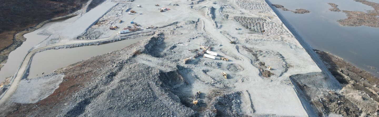 Aerial view of a large open-pit mine site surrounded by rocky, barren terrain and water bodies, with construction vehicles and equipment scattered across the excavation area.