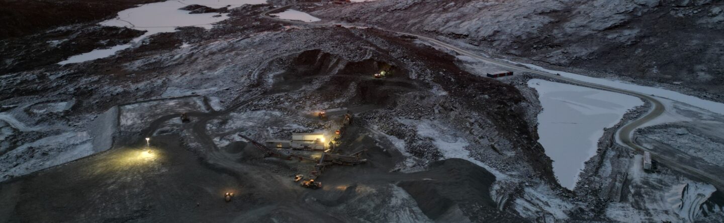 Aerial view of a mining site at dawn, with snowy mountains and frozen lakes in the background. Machinery with headlights is working on the rocky terrain under a colorful sunrise sky.