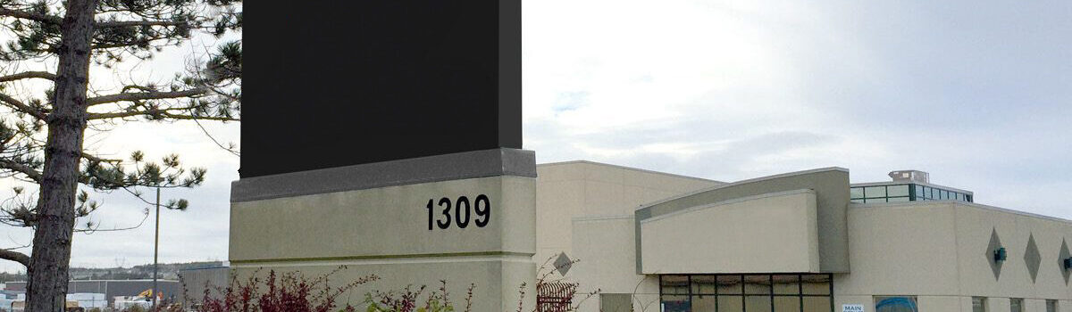 A large blank black sign displays the number 1309 in front of a beige commercial building, with a tree and overcast sky in the background.