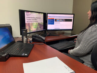 A person sitting at a desk with a laptop and two monitors; one monitor displays a presentation titled “Understanding Historical Treaties and Métis Assertion of Rights,” and the other shows a website.