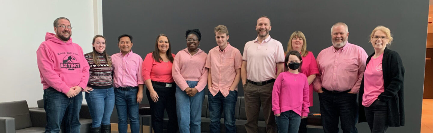 A group of twelve people stand indoors in front of a dark gray wall, all wearing pink shirts or sweaters, smiling and posing for a group photo.