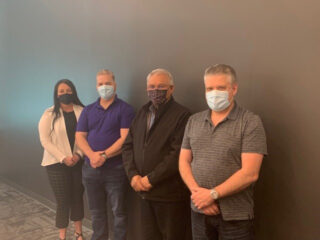 Four people wearing face masks stand in front of a wall. Three men and one woman stand side by side, posing for the photo in an office setting.