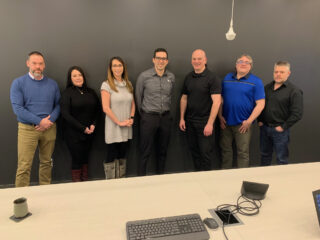 Seven people stand in a row smiling in front of a dark wall with the “Pennecon” logo. A desk with a keyboard and phone is in the foreground. The group is dressed in business casual attire.